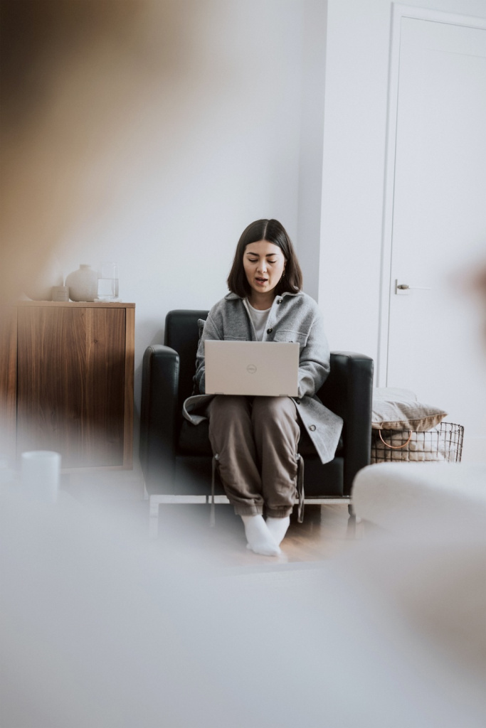 A woman sits on a black armchair working on a laptop. She is wearing a gray coat and khaki pants. The room is softly lit, featuring a wooden cabinet with a teapot on it and a white door in the background.